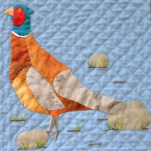 May include: A colorful patchwork quilt block featuring a pheasant with a red head and a blue neck, standing on a blue background with brown rocks and green grass.