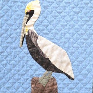 May include: A brown and white pelican with a yellow head and a long orange beak stands on a brown log against a blue quilted background.