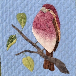 May include: A pink and brown bird perched on a branch with green leaves. The bird is a quilted fabric applique on a blue quilted background.