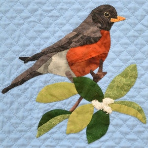 May include: A textile artwork depicting a robin bird applique on a branch with leaves and small white flowers. The bird has a grey back, orange breast, and black head. The background is a light blue quilted fabric with a diamond pattern.