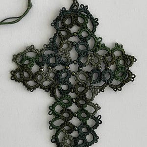 Puede incluir: Colgante de cruz calada hecho a mano en tonos verdes y grises. El intrincado diseño presenta una serie de bucles interconectados y pequeñas cuentas. El colgante cuelga de un cordón verde a juego, creando un accesorio único y detallado.