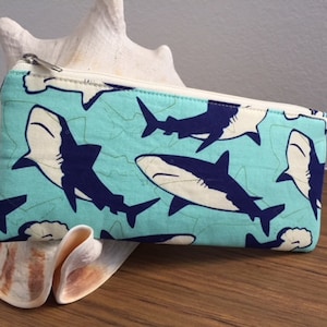 May include: A blue and white zippered pouch with a shark print. The pouch is made of fabric and has a zipper closure.