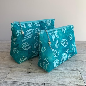 May include: Two turquoise blue zippered pouches with a white seashell and starfish pattern. The pouches are made of fabric and have a zipper closure. The smaller pouch is in front of the larger pouch.