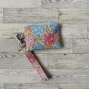 May include: A patchwork floral print wristlet wallet with a light blue zipper and a silver-tone clasp. The wrist strap and wallet feature a mix of pink, blue, and orange floral patterns. The wallet measures approximately 5 inches wide and 3.5 inches tall.