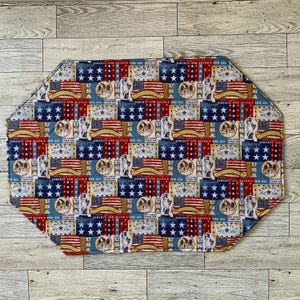 May include: An octagonal placemat with a patriotic design. The pattern features American flags, stars, and the text "Liberty and Justice for All" in red, white, and blue. The placemat is made of fabric.
