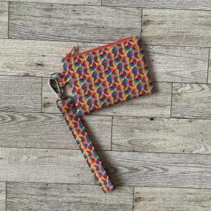 May include: A small zipped pouch and wrist strap, both patterned with a vibrant geometric design. The pattern features repeating multi-coloured triangles in red, yellow, blue, and green. The pouch has a silver clasp and a red zip.