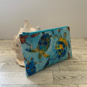 May include: A turquoise zippered pouch with a blue background and a pattern of colorful sea turtles. The pouch has a silver charm attached to the zipper.