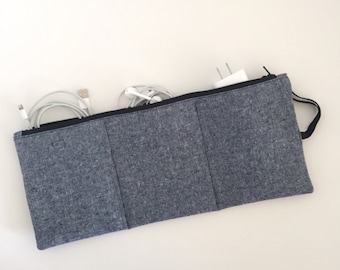 May include: A rectangular charcoal gray fabric pouch with a black zipper closure and a small side loop. The pouch features three vertical stitched sections on the front and contains white electronic charging cables and a white power adapter.