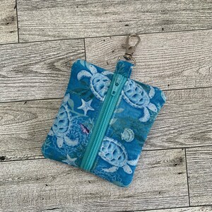 May include: A small blue zippered pouch with a sea turtle print. The pouch has a silver zipper and a silver clip attached to the top.