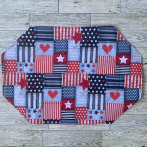May include: A patriotic placemat with a patchwork design featuring red, white, and blue fabrics. The design includes stars, stripes, and hearts. The placemat is approximately 18 inches long and 12 inches wide.