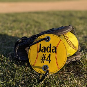 May include: Yellow softball with black stitching and a yellow personalized tag with black stitching that says "Jada #4". The tag is attached to a black leather softball glove.