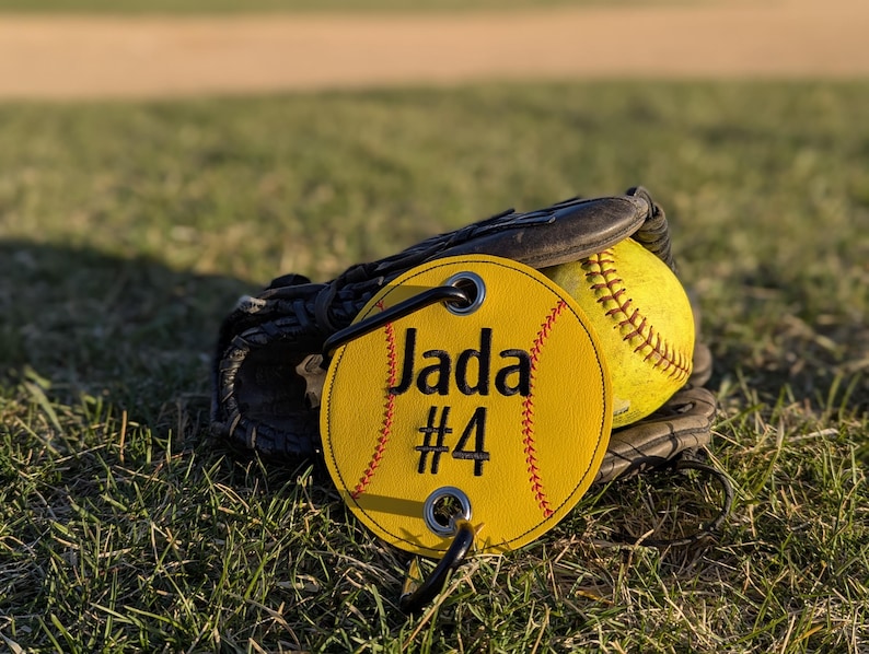 May include: Yellow softball with black stitching and a yellow personalized tag with black stitching that says "Jada #4". The tag is attached to a black leather softball glove.