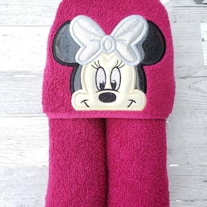 May include: A pink hooded towel with a black and white embroidered Minnie Mouse design. The bow on Minnie Mouse is silver.