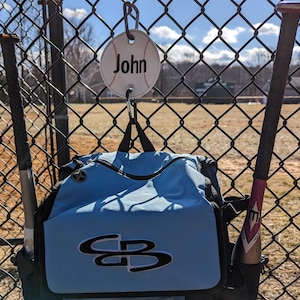 May include: A blue baseball bag with a white and black logo hanging on a chain link fence. A baseball bat is leaning against the bag. A white baseball with the name "John" written on it is hanging from the chain link fence.