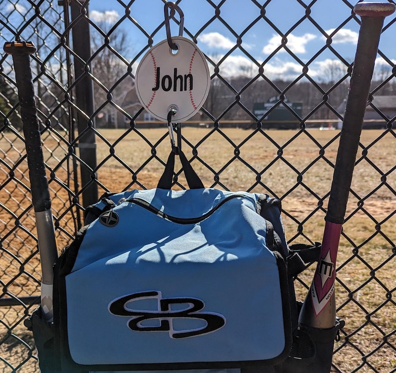 May include: A blue baseball bag with a white and black logo hanging on a chain link fence. A baseball bat is leaning against the bag. A white baseball with the name "John" written on it is hanging from the chain link fence.