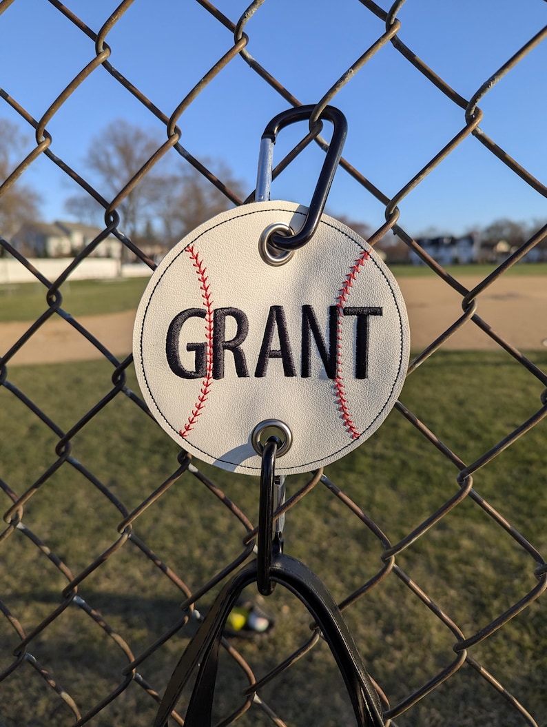 May include: White baseball-shaped tag with red stitching and the name "GRANT" embroidered in black. The tag is attached to a black carabiner clip.