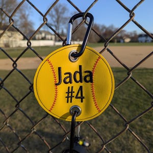 May include: Yellow softball-shaped keychain with the name "Jada" and the number "#4" embroidered in black thread. The keychain has a black carabiner clip.