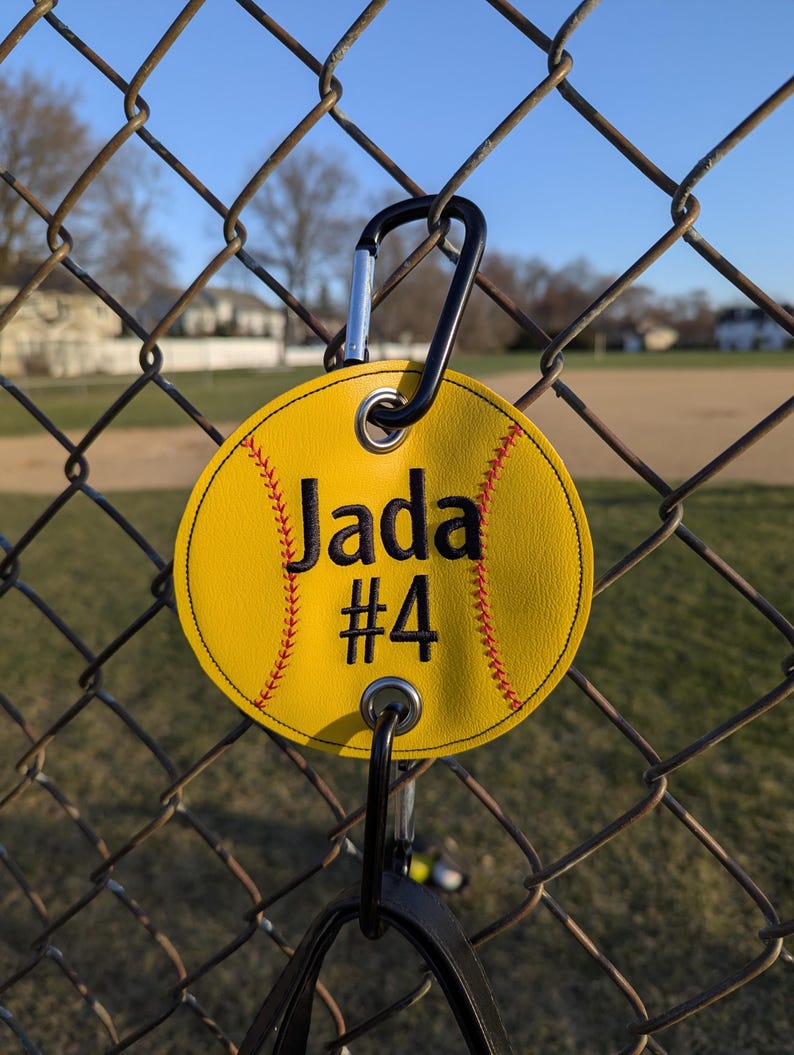 May include: Yellow softball-shaped keychain with the name "Jada" and the number "#4" embroidered in black thread. The keychain has a black carabiner clip.