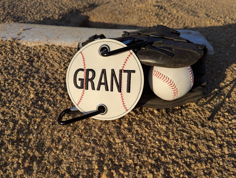 May include: White baseball-shaped tag with black stitching and the name "GRANT" embroidered in black. The tag has a black carabiner clip attached.