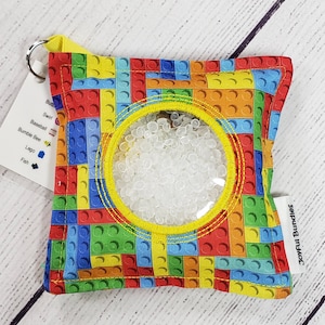 May include: A colorful sensory toy with a clear window filled with white beads. The fabric is patterned with colorful Lego bricks and has a yellow drawstring. The tag reads "Joyful Bundles".