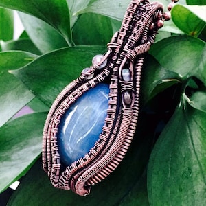 Puede incluir: Un colgante envuelto en alambre de cobre con una piedra preciosa de ágata azul. El colgante cuelga de una cadena de cobre.