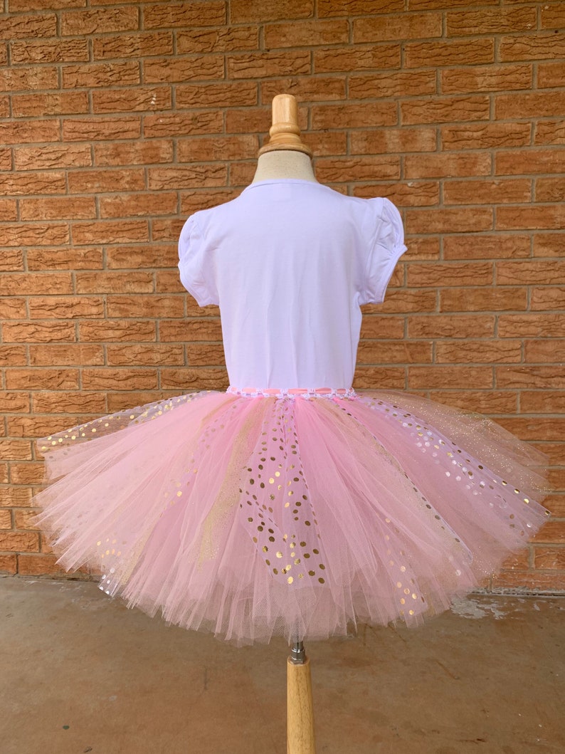 May include: A pink tulle tutu skirt with gold glitter polka dots. The skirt is on a white mannequin.