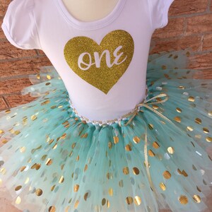 May include: A white short-sleeved shirt with a gold glitter heart that says "one" on it. The shirt is paired with a mint green tulle tutu with gold polka dots.