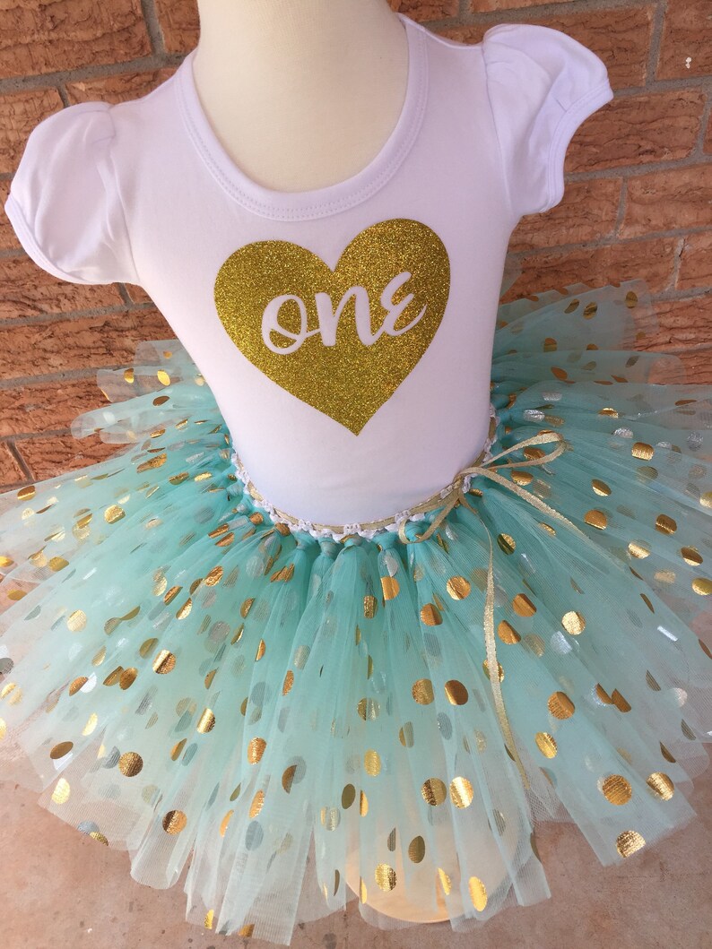 May include: A white short-sleeved shirt with a gold glitter heart that says "one" on it. The shirt is paired with a mint green tulle tutu with gold polka dots.