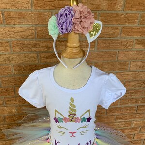 May include: A white t-shirt with a unicorn design featuring a gold glitter horn, pink and purple flowers, and the number 7. The shirt is paired with a rainbow tulle tutu with gold glitter polka dots and a gold ribbon. The name "Katelyn" is written in purple glitter on the shirt.
