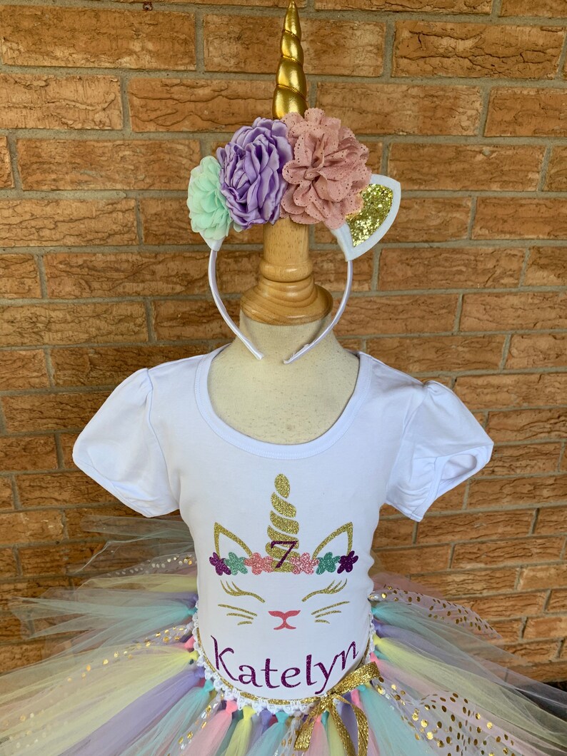 May include: A white t-shirt with a unicorn design featuring a gold glitter horn, pink and purple flowers, and the number 7. The shirt is paired with a rainbow tulle tutu with gold glitter polka dots and a gold ribbon. The name "Katelyn" is written in purple glitter on the shirt.