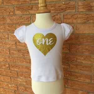 May include: White short-sleeved shirt with a gold glitter heart that says "one" on it.