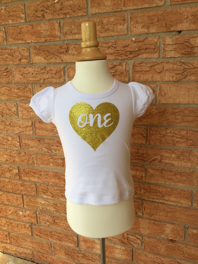 May include: White short-sleeved shirt with a gold glitter heart that says "one" on it.