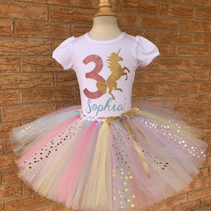 May include: A white t-shirt with a gold glitter unicorn and the number 3 in pink glitter. The shirt says "Sophia" underneath the unicorn. The shirt is paired with a multi-colored tulle tutu with gold glitter polka dots.