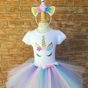 Peut inclure: Une tenue de tutu de licorne blanche pour filles, comprenant un t-shirt blanc avec un visage de licorne paillet&eacute;, une jupe tutu en tulle dans les tons rose, violet et bleu, et un serre-t&ecirc;te de licorne blanc avec une corne dor&eacute;e et des fleurs roses, violettes et bleues.