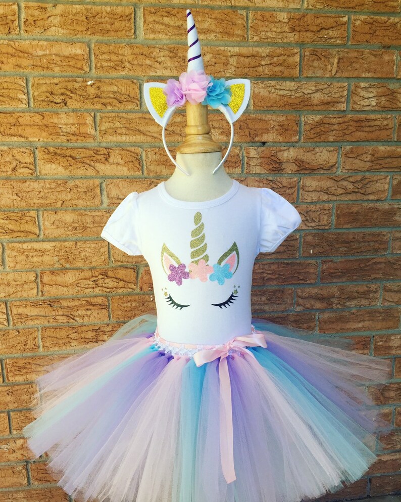 Peut inclure: Une tenue de tutu de licorne blanche pour filles, comprenant un t-shirt blanc avec un visage de licorne paillet&eacute;, une jupe tutu en tulle dans les tons rose, violet et bleu, et un serre-t&ecirc;te de licorne blanc avec une corne dor&eacute;e et des fleurs roses, violettes et bleues.