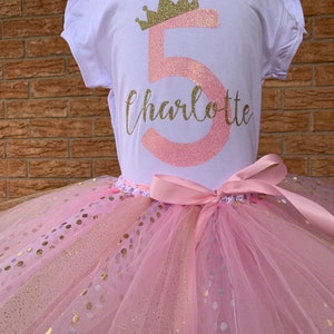 May include: A pink and gold tutu with gold polka dots. A white t-shirt with a pink number 5 and a gold crown with the name Charlotte written below.