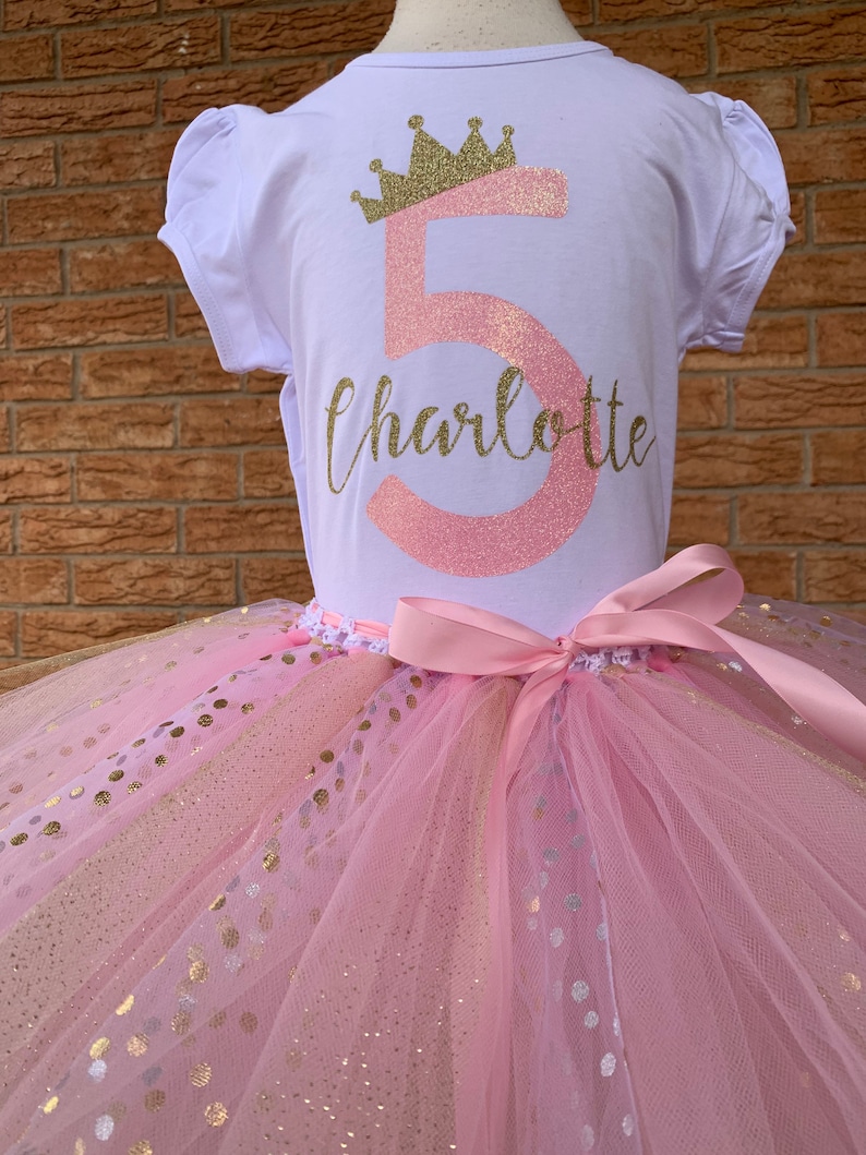 May include: A pink and gold tutu with gold polka dots. A white t-shirt with a pink number 5 and a gold crown with the name Charlotte written below.