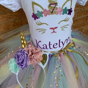 May include: A white t-shirt with a gold glitter unicorn design, a pink and purple flower crown, and the name "Katelyn" written in purple glitter. The shirt is paired with a rainbow tulle tutu with gold glitter dots and a white unicorn headband with gold glitter ears and a gold horn.
