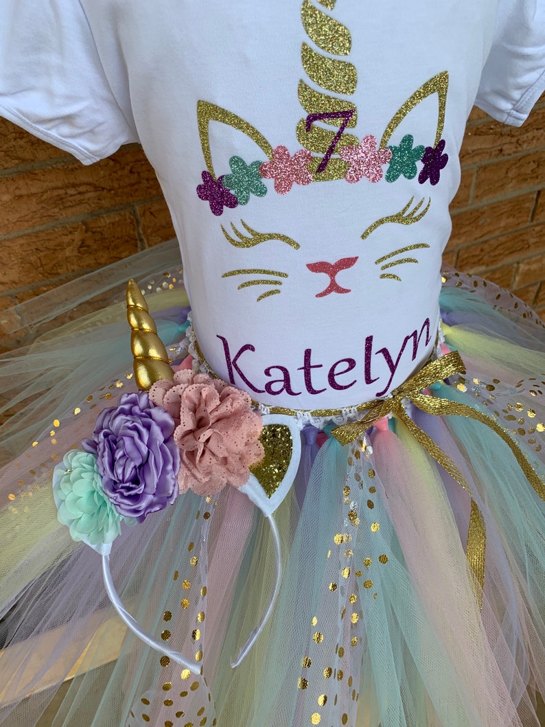 May include: A white t-shirt with a gold glitter unicorn design, a pink and purple flower crown, and the name "Katelyn" written in purple glitter. The shirt is paired with a rainbow tulle tutu with gold glitter dots and a white unicorn headband with gold glitter ears and a gold horn.