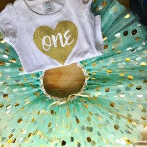 May include: A white onesie with a gold glitter heart that says "one" on it. The onesie is paired with a turquoise tulle tutu with gold glitter polka dots.