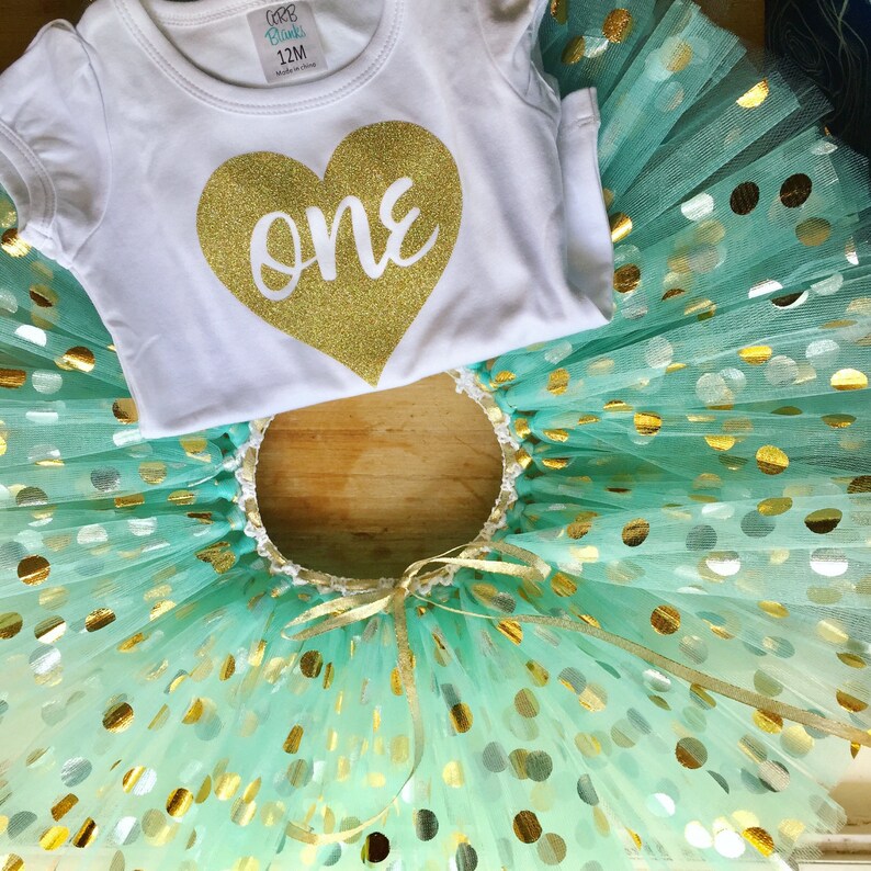 May include: A white onesie with a gold glitter heart that says "one" on it. The onesie is paired with a turquoise tulle tutu with gold glitter polka dots.
