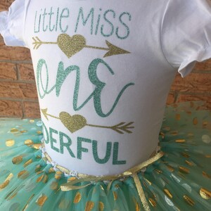 May include: A white shirt with the text "Little Miss One Derful" in teal and gold glitter. The shirt is paired with a teal tulle skirt with gold polka dots.