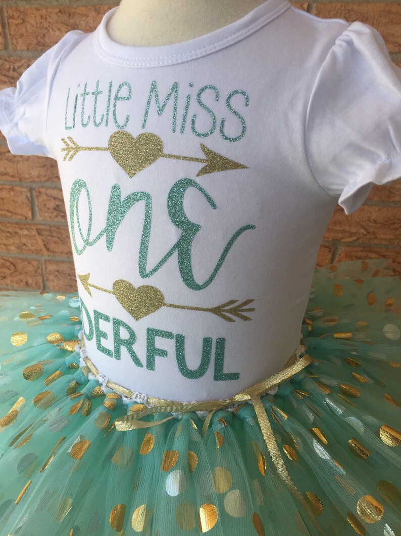 May include: A white shirt with the text "Little Miss One Derful" in teal and gold glitter. The shirt is paired with a teal tulle skirt with gold polka dots.