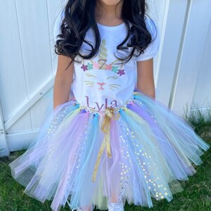 May include: A young girl wearing a white t-shirt with a unicorn design and the name "Lyla" on it. She is also wearing a rainbow tulle tutu with gold glitter accents.