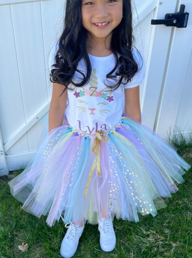 May include: A young girl wearing a white t-shirt with a unicorn design and the name "Lyla" on it. She is also wearing a rainbow tulle tutu with gold glitter accents.