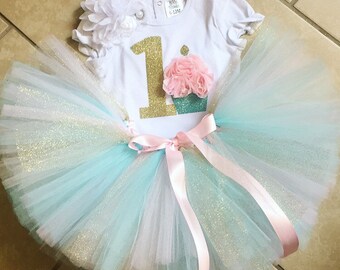 May include: A white onesie with a gold glitter number one and a cupcake graphic. The onesie is paired with a turquoise and white tulle tutu with a pink satin ribbon.