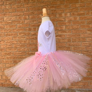 May include: A pink tulle tutu skirt with gold glitter dots. The skirt is worn on a white mannequin with a white short-sleeved shirt that says "One" in gold glitter.