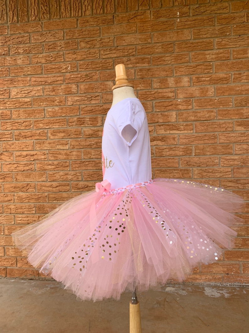 May include: A pink tulle tutu skirt with gold glitter dots. The skirt is worn on a white mannequin with a white short-sleeved shirt that says "One" in gold glitter.