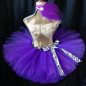 May include: Purple tulle tutu skirt with a zebra print ribbon and a purple bow. The tutu has glitter throughout.