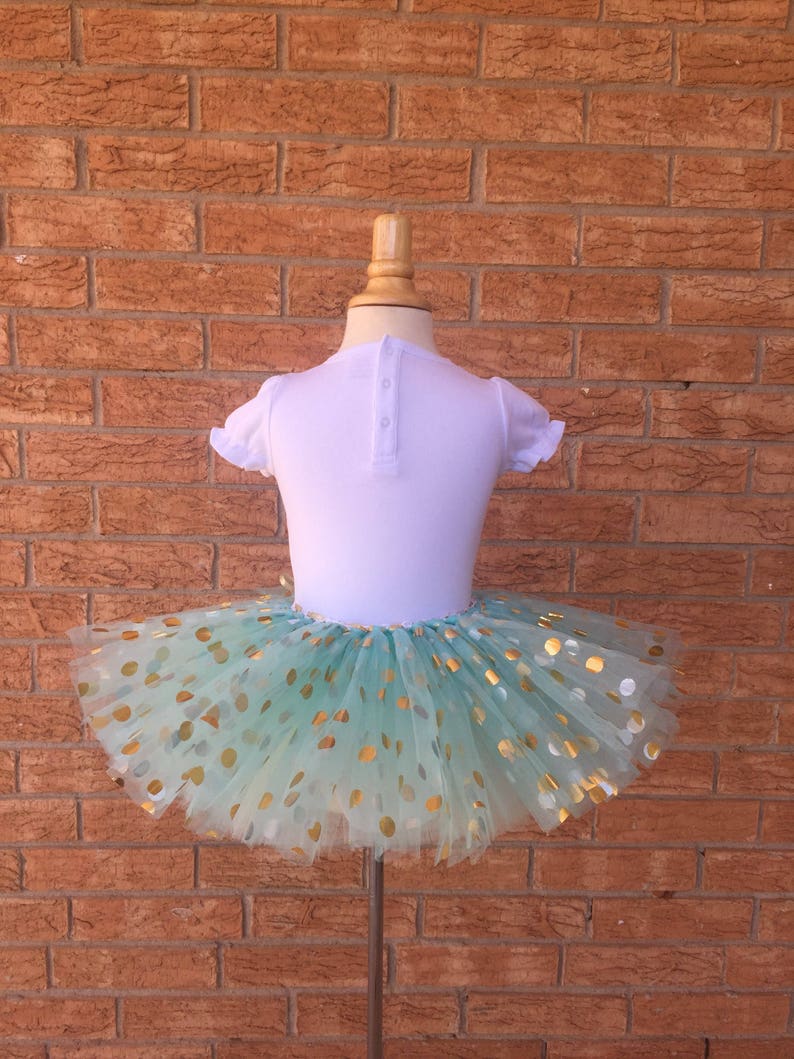 May include: A mint green tulle tutu skirt with gold polka dots. The skirt is on a white mannequin.